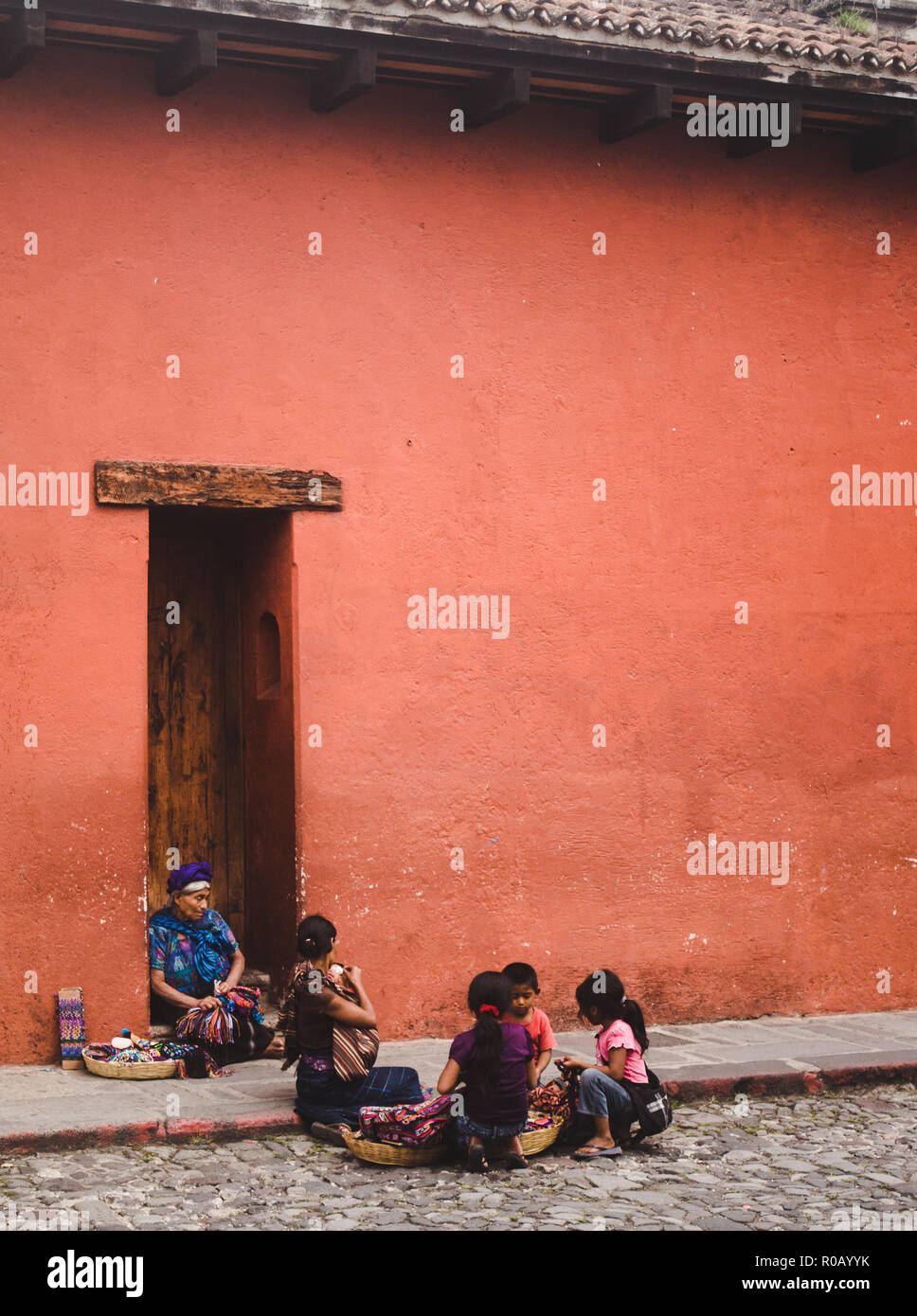 Matriarchin der Guatemaltekischen Familie im traditionellen Maya Kleid verkauft Schmuck & Tücher von Körben auf der Straße neben ihrer Familie in Antigua Guatemala Stockfoto