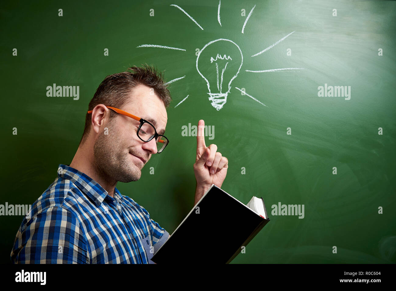 Eine junge Zerzausten Mann mit Brille ist ein Buch lesen, kommt eine Idee auf dem Hintergrund einer Tafel mit Kreide Glühbirne zu kümmern. Stockfoto