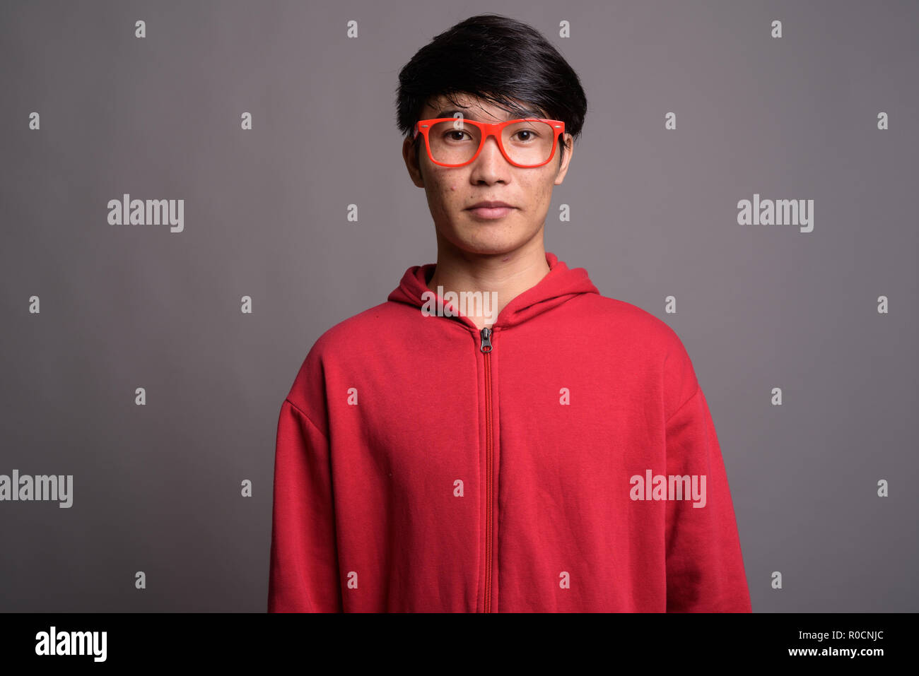 Jungen asiatischen Mann mit roten Mantel mit Brillen gegen Grau Stockfoto