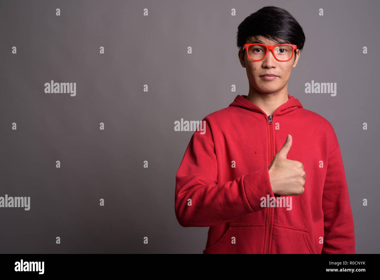 Jungen asiatischen Mann mit roten Mantel mit Brillen gegen Grau Stockfoto