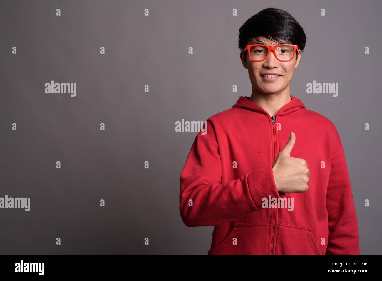 Jungen asiatischen Mann mit roten Mantel mit Brillen gegen Grau Stockfoto