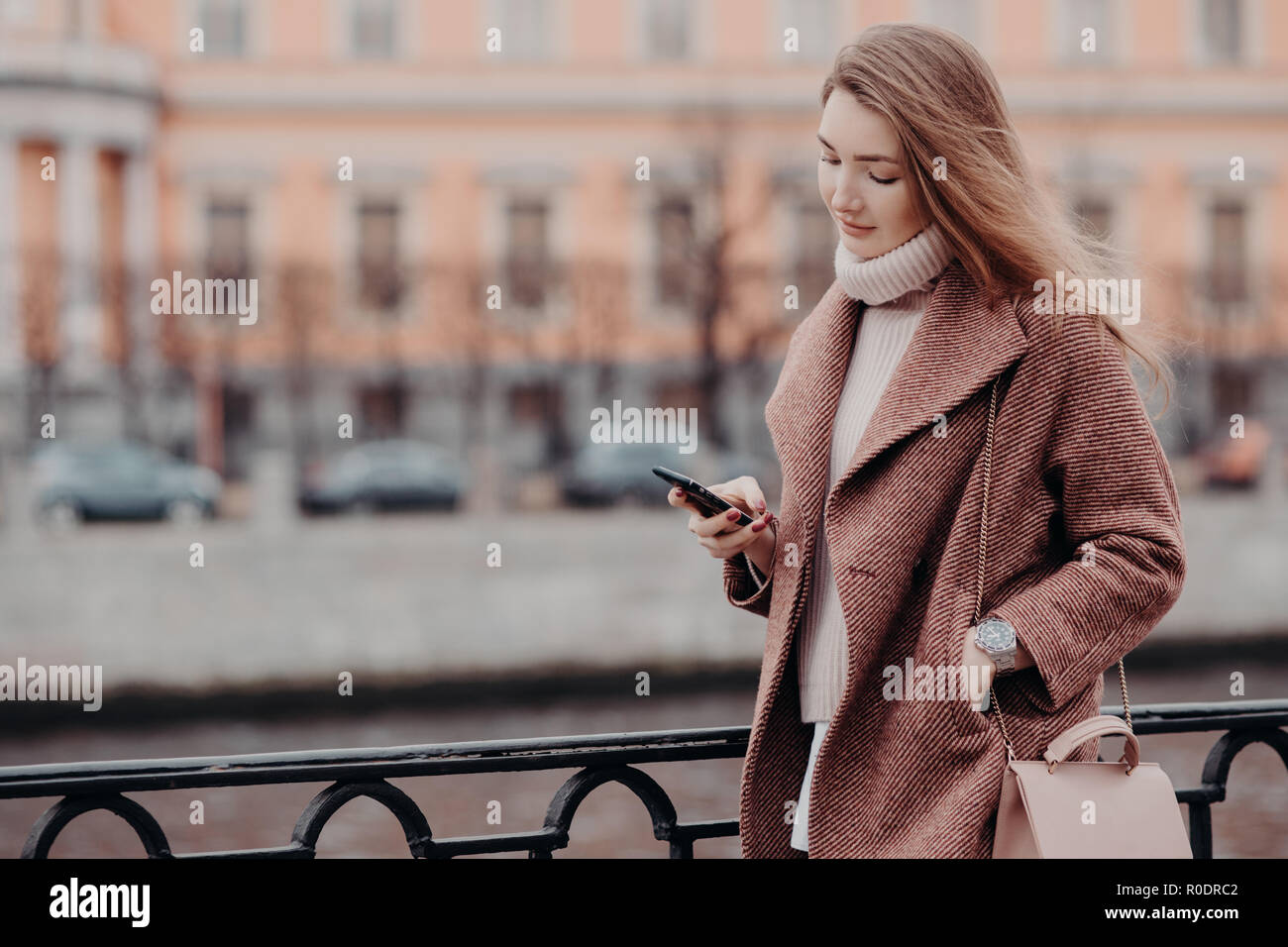 Schöne Frau in warmer Mantel, hält ein modernes Handy, Nachrichten in sozialen Netzwerken, dem kostenfreien WLAN verbunden, in der Stadt zu Fuß, street style, verschwommenes backgr Stockfoto