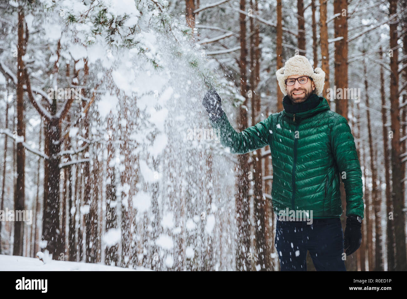 Portrait von fröhlichen stattliche männliche trägt Pelzkappe mit Ohr Klappe und gelbe und grüne Jacke aus, wirft Schnee in der Luft, genießt, freie Zeit in die schöne Stockfoto