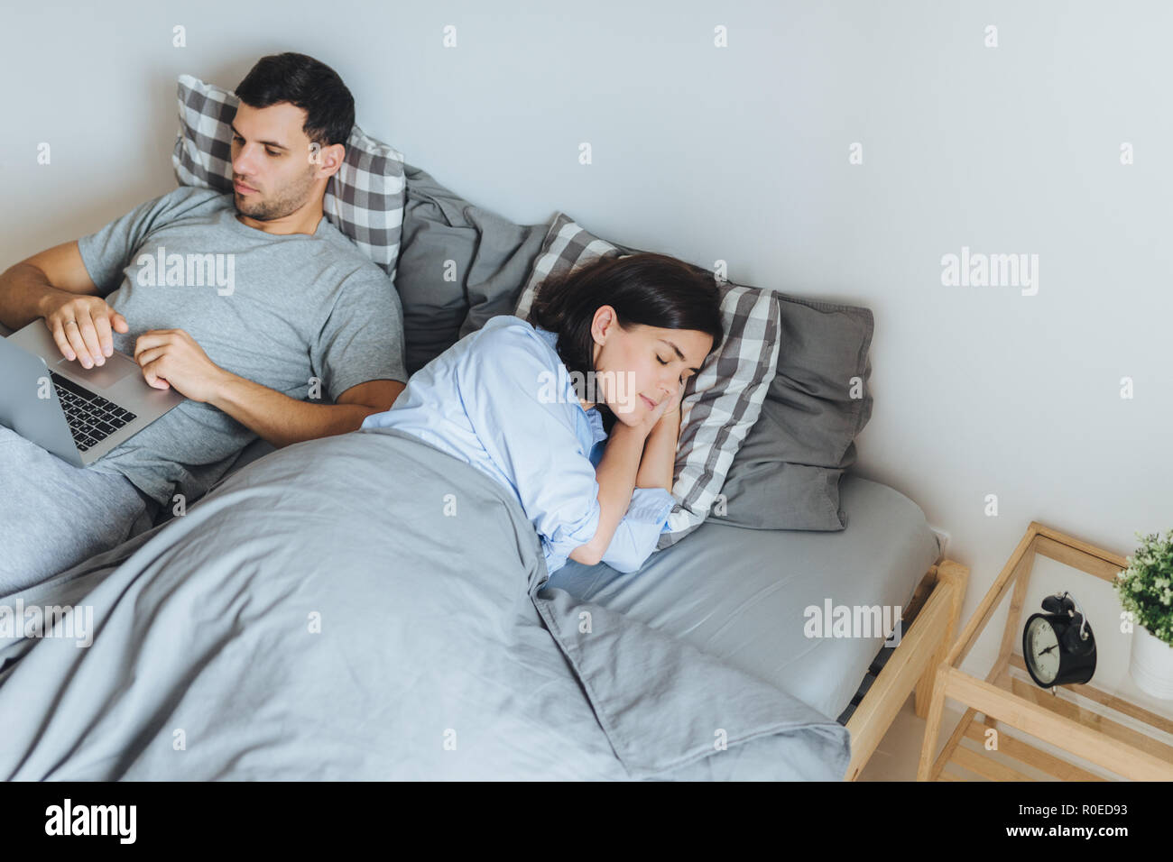 Hübsche Frau schläft im Bett, sieht angenehme Träume, während ihr Mann am Laptop arbeitet, der Bericht bereitet, versucht nicht seine Frau Schlaf zu unterbrechen. Stockfoto