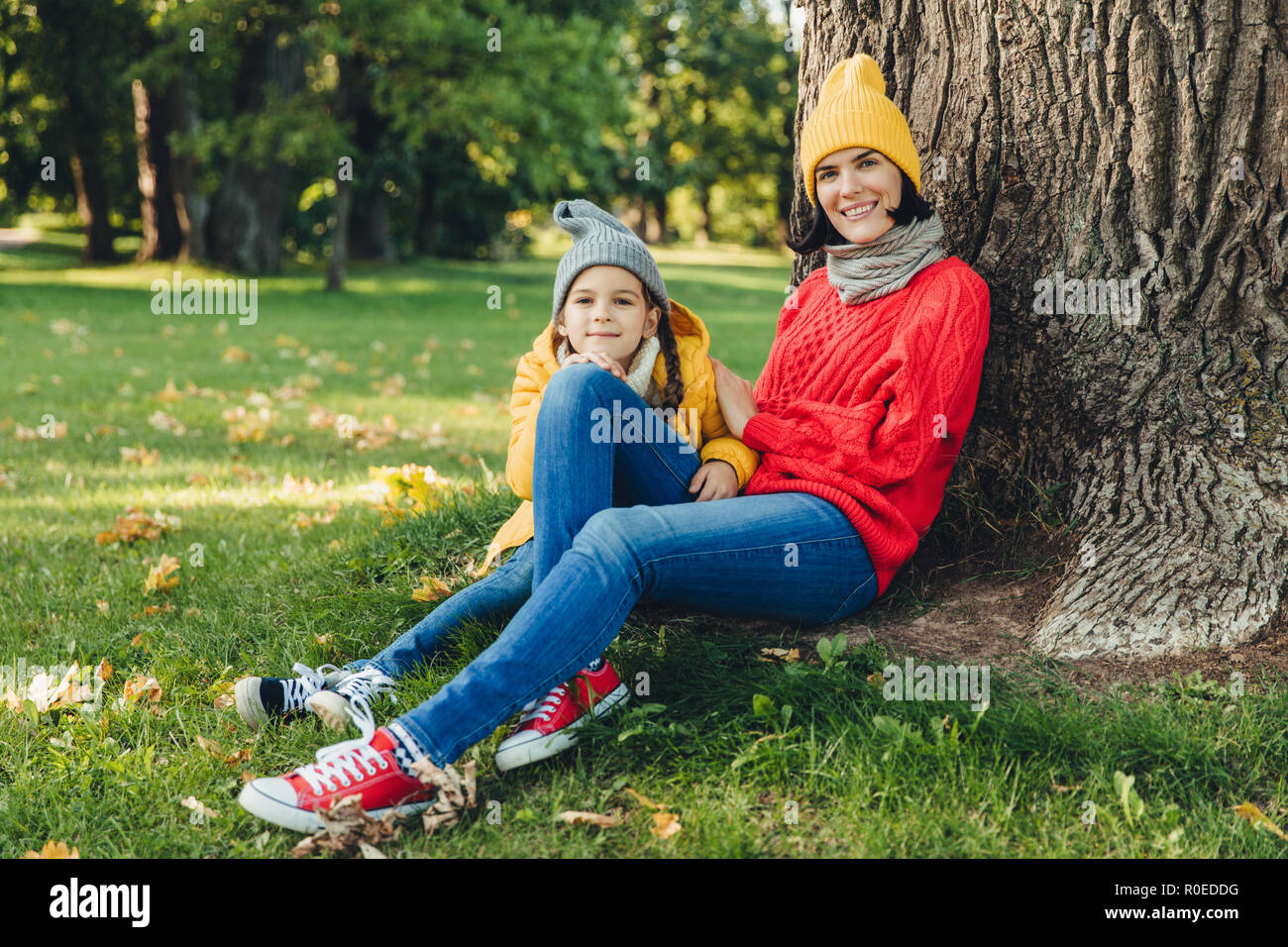 Outdoor Portrait von schönen Frauen in Mütze und warme lose Pullover bekleidet Wochenenden verbringen zusammen mit ihrer kleinen Tochter, sitzen auf grünem Gras n Stockfoto