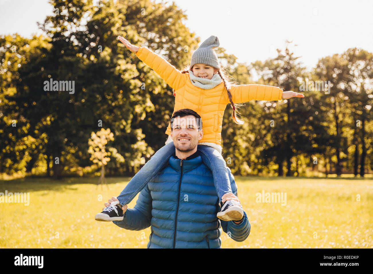 Stattliche Vater trägt anorack piggyback seinen wenig lustig Mädchen, schlendern Sie gemeinsam im Freien, die Sonne zu genießen. Attraktive Vati und seine Tochter, die r Stockfoto