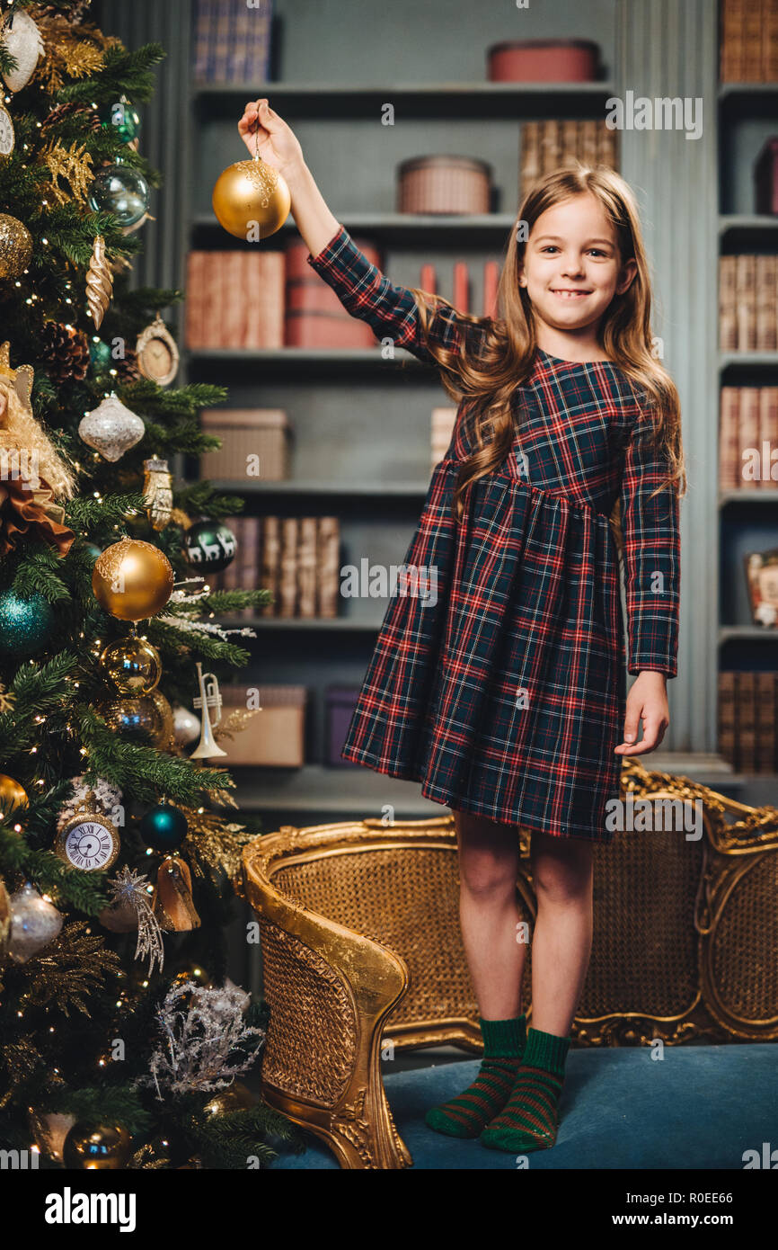 Hübsches Mädchen mit charmanten Lächeln steht auf Sessel, schmückt allein Neues Jahr Baum, hat gute Laune, fühlt sich stolz, es selbst zu tun. Kleines Kind bereitet Stockfoto