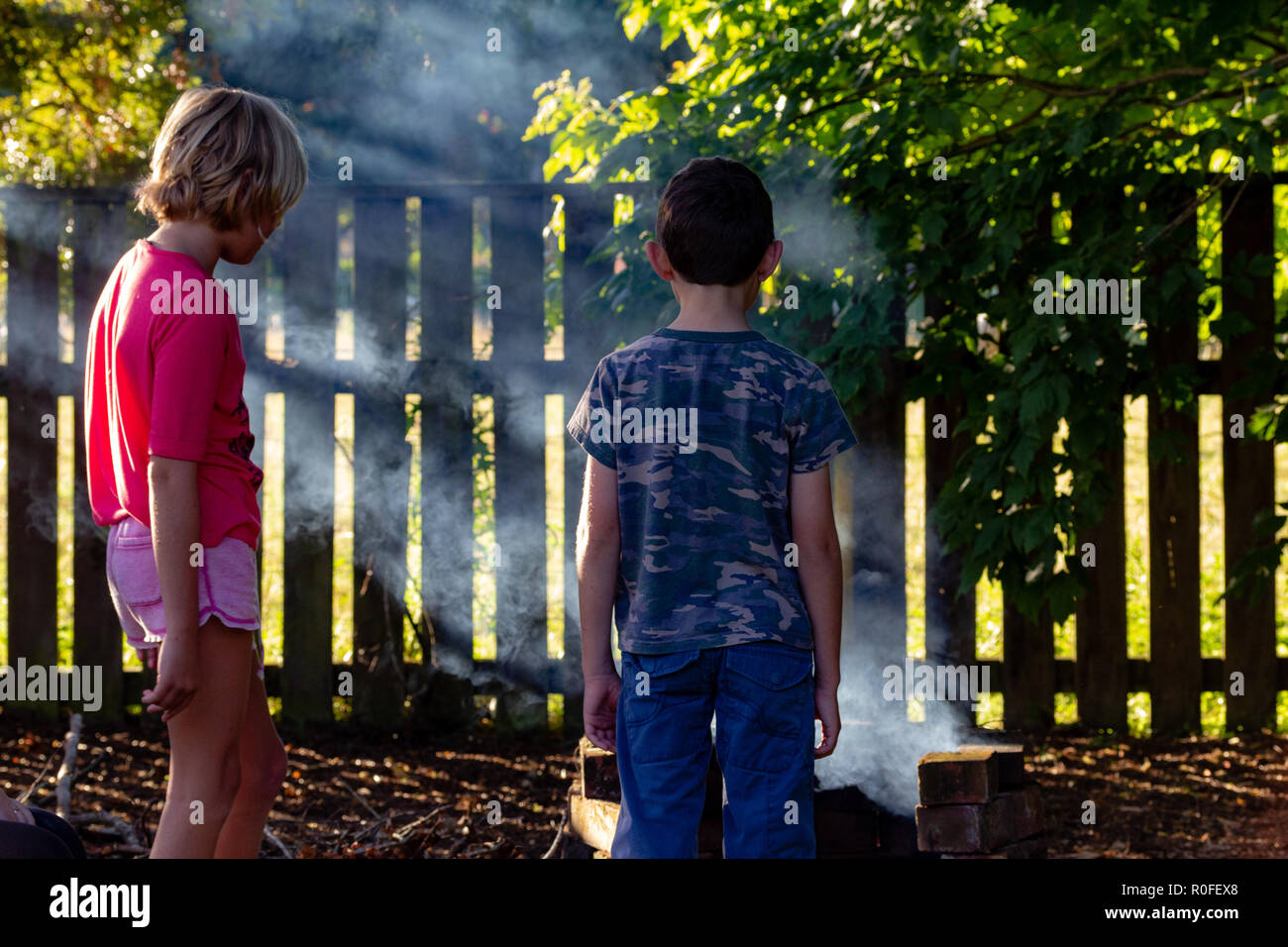 Authentisches Bild von zwei Kindern draußen im Garten in Rauch auf Feuer, am späten Nachmittag Sonne. Kinder aus Geräten und in der freien Natur. Stockfoto