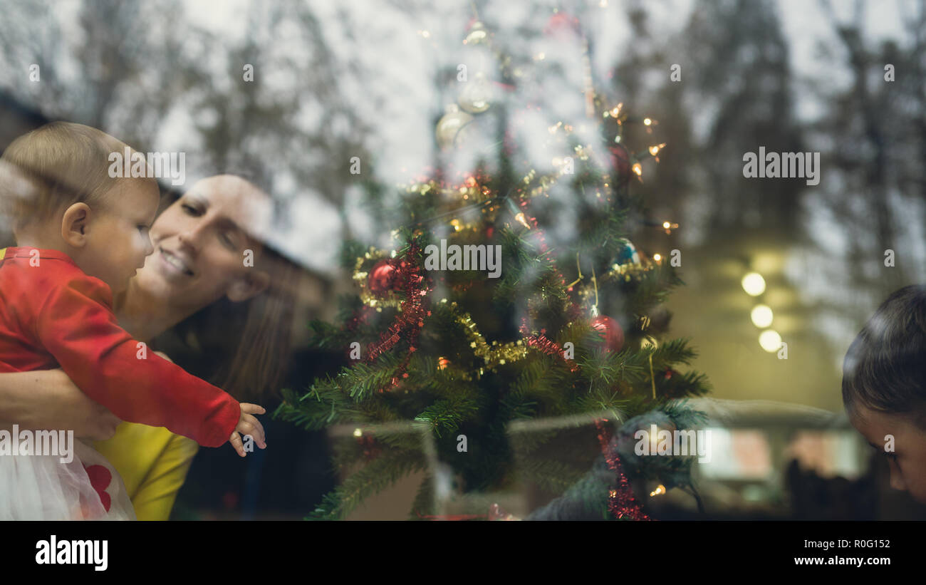 Retro Bild einer glücklichen jungen Mutter ihr Baby Mädchen Weihnachtsbaum bewundern, die auch heute noch von ihren Toddler Sohn eingerichtet werden. Blick durch ein Wi Stockfoto
