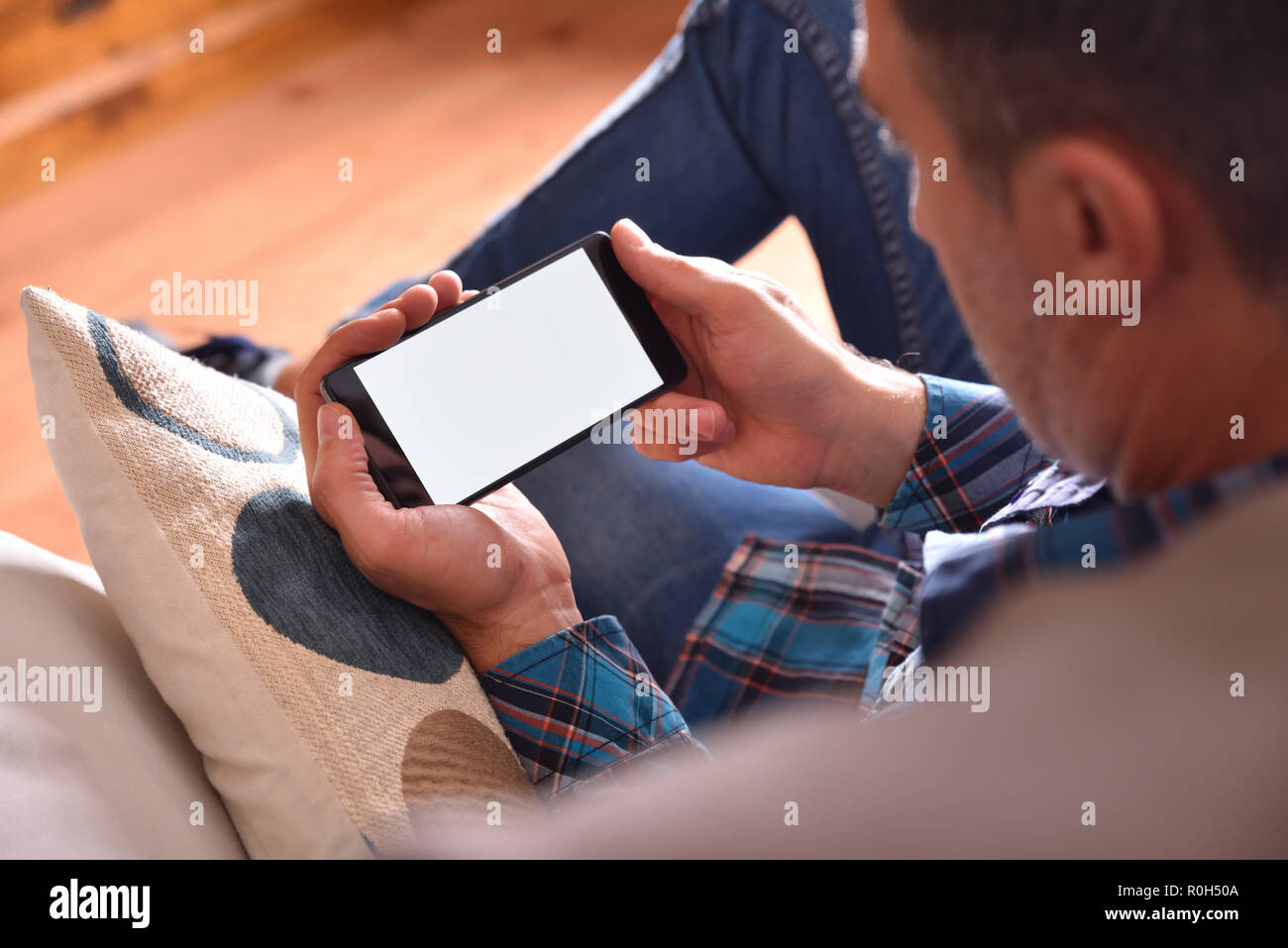 Erwachsenen Mann mit blauem Hemd und Jeans sitzt auf einem Sofa, multimediale Inhalte auf einem Smartphone. Aussichtspunkt angehoben Stockfoto