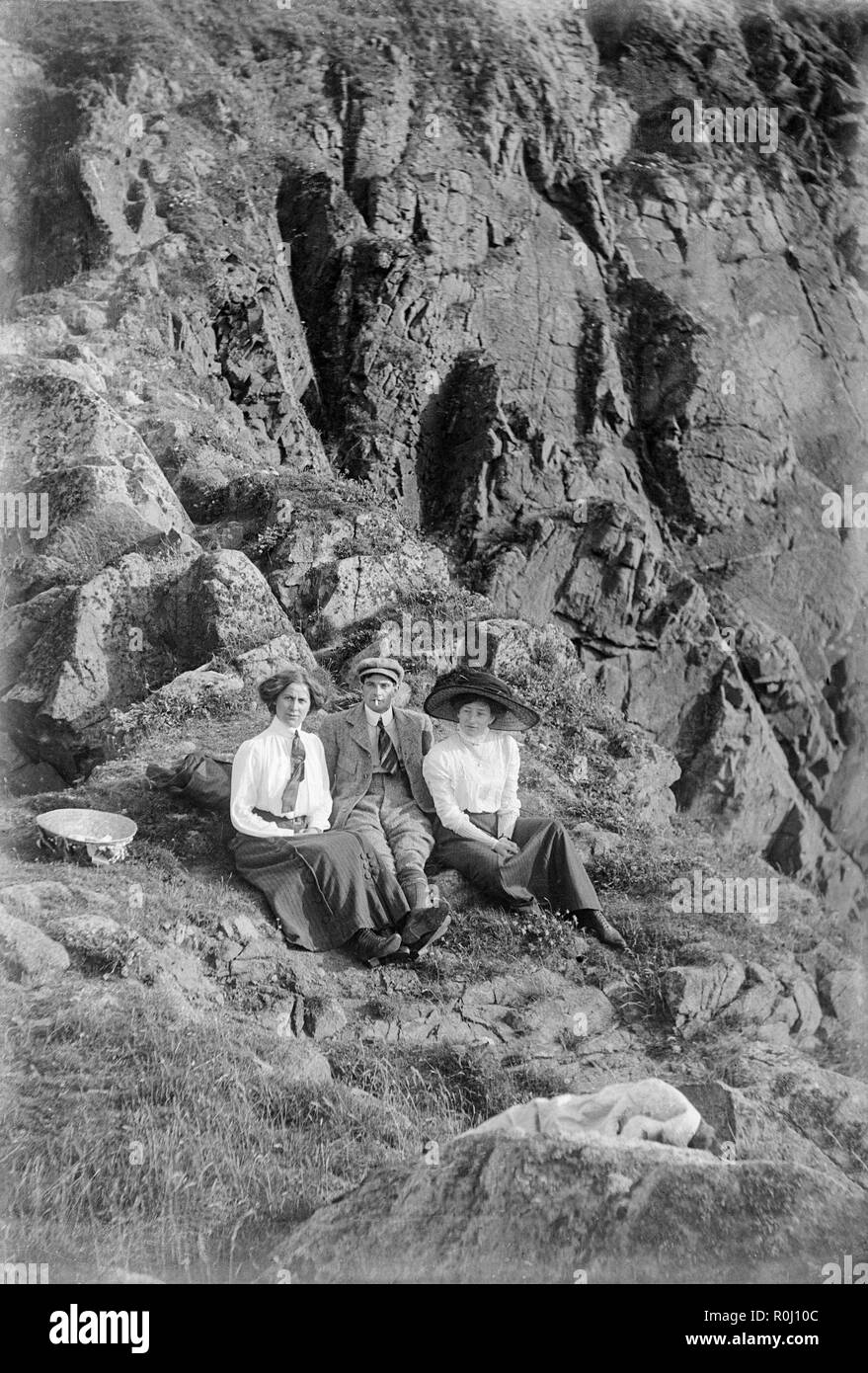 Edwardian schwarz-weiß Foto, ein junger Mann und zwei junge Frauen, sitzen auf dem Boden einer felsigen Klippe, irgendwo in England. Alle tragen Kleidung in guter Qualität der Ära. Eine Frau trägt einen sehr großen Hut, typisch für die edwardianische Zeit. Stockfoto