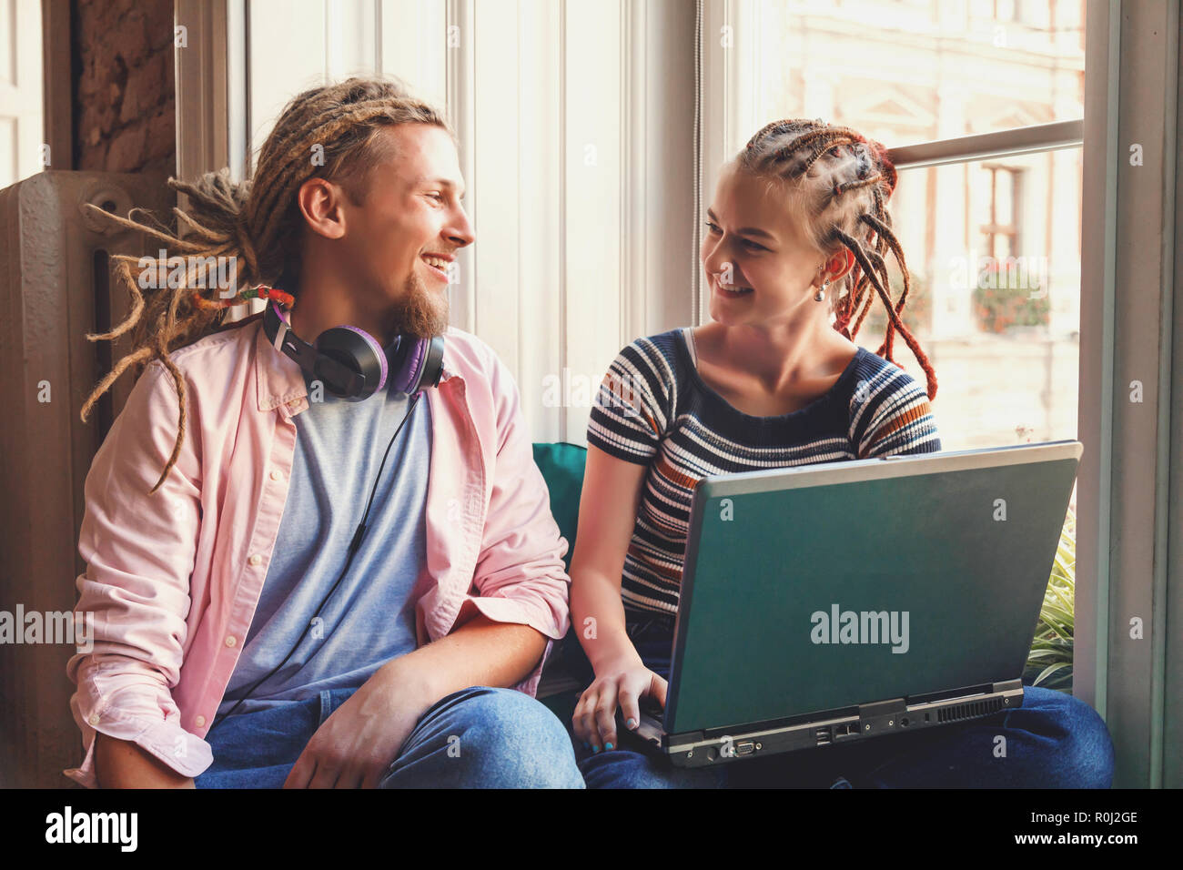 Zwei hipster beste Freunde mit dreads sitzen in der Nähe der Fenster und und plaudern miteinander, Mädchen, dass Laptop und zeigt Bilder zu Ihrem Freund Stockfoto