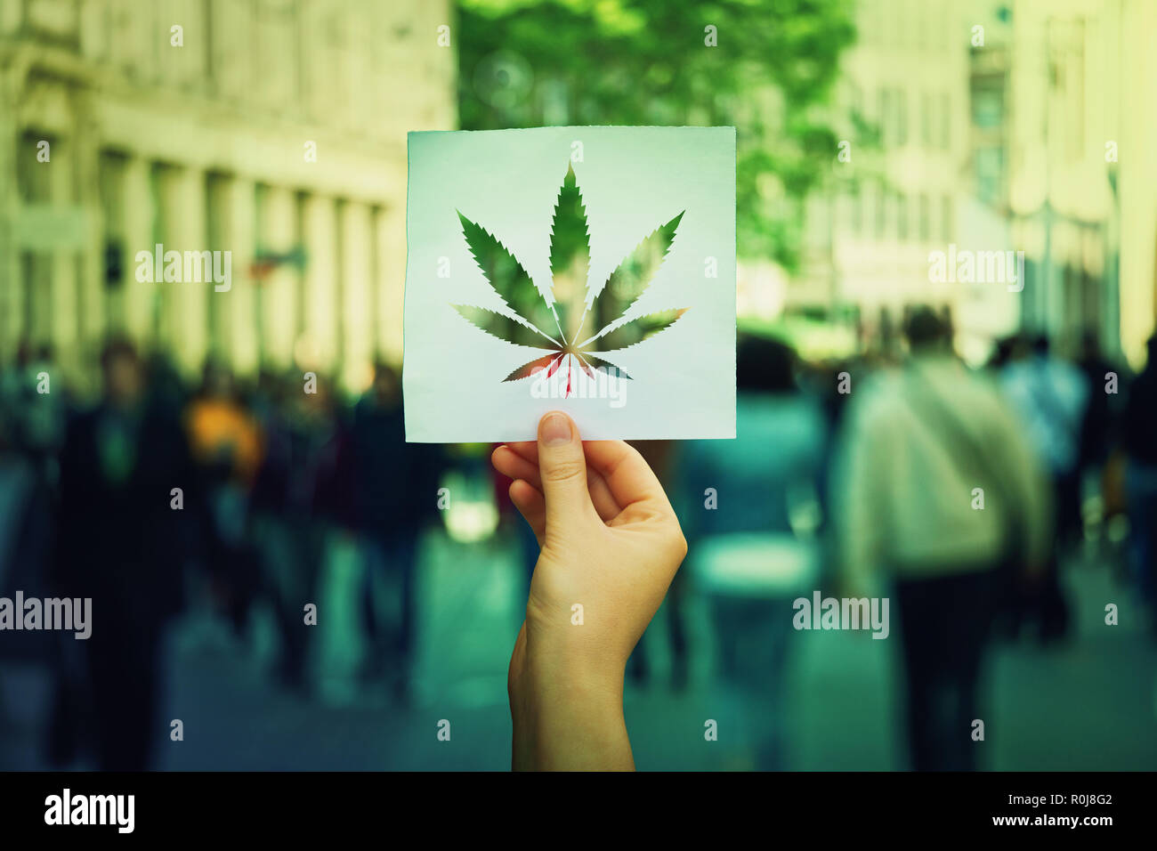 Hand hält ein Blatt Papier mit Marihuana Blatt Symbol über einem überfüllten Straße Hintergrund. Legalisierung von Cannabis als Medikament. CBD Heilung sozialen issu Stockfoto