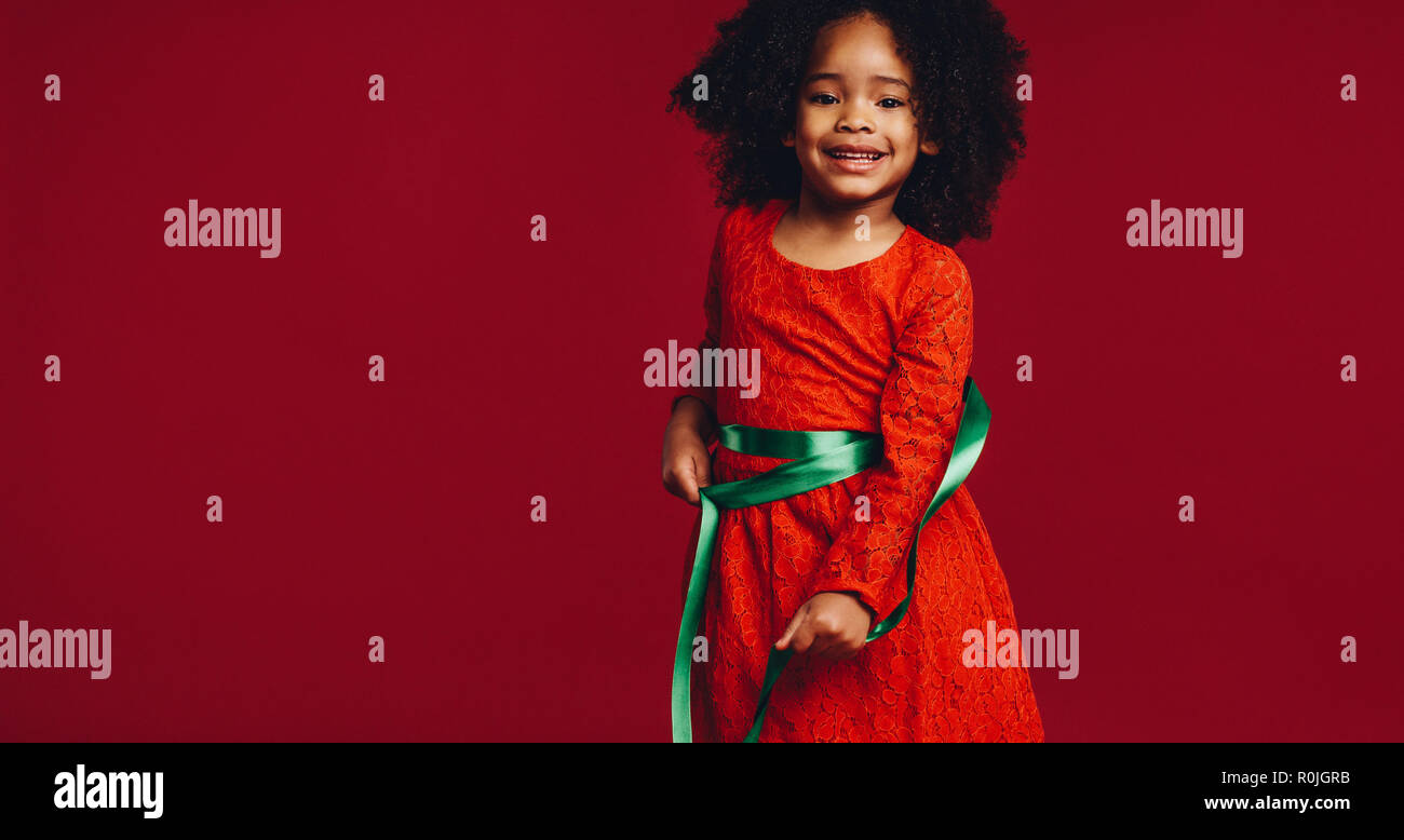 Lächelnd Kind spielt mit einem Satinband um ihre Taille. Afro-amerikanischen Jungen auf rotem Hintergrund spielen mit einem dekorativen Band stehen. Stockfoto