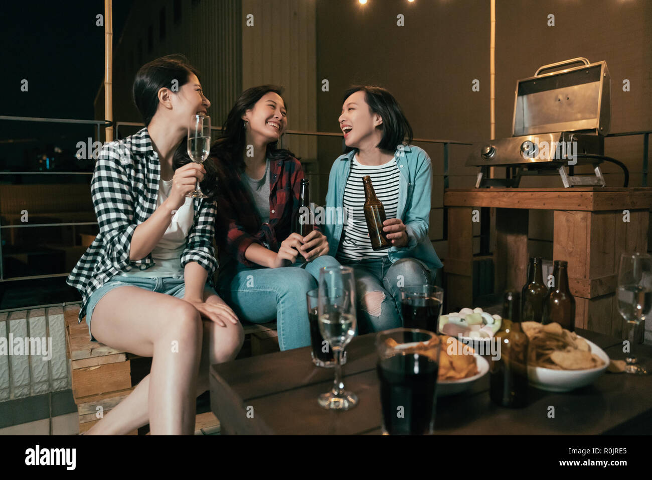 Asiatische Mädchen Spaß Lachen auf dem Dach Partei. Junge Damen chatten trinken Alkoholen sitzen draussen in der Nacht. Grill Event mit Bier und gewinnen Stockfoto
