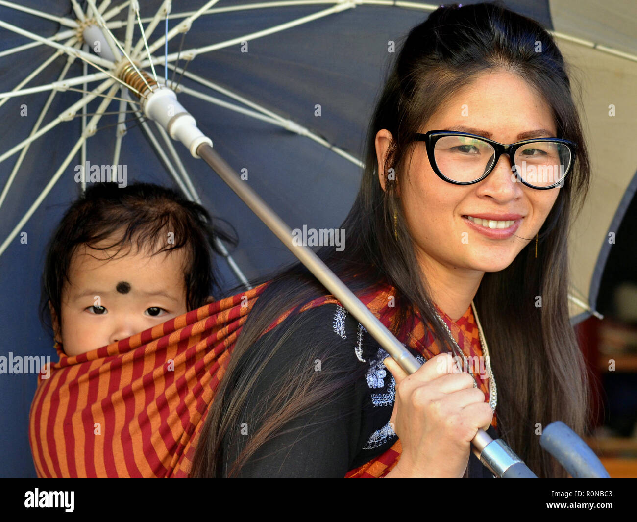 Jungen nordöstlichen indischen Monpa Frau mit modernen Brillen trägt ihr baby boy in einem traditionellen Babytragetuch unter einen Schirm auf dem Rücken. Stockfoto