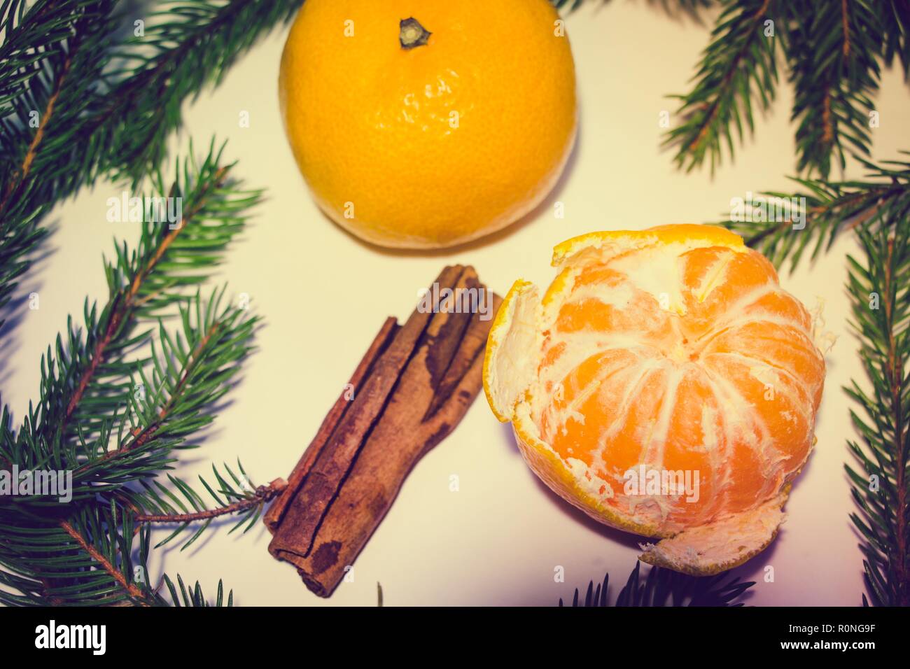 Zwei Mandarinen, von denen einer geschält wird, auf einem weißen Hintergrund. Um die Mandarinen sind Tannenzweigen und ein paar Stöcke von duftenden Zimt. Stockfoto