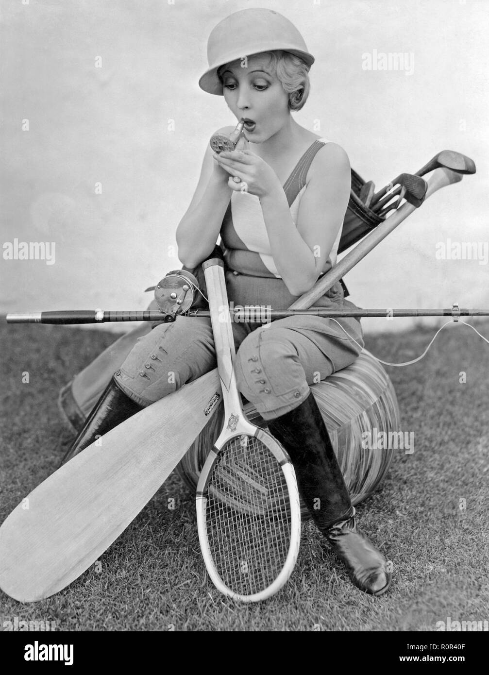 Bessie Love, 1898-1986. Die amerikanische Schauspielerin hier dargestellt mit einem Lippenstift und einen kleinen Taschenspiegel. Sie ist durch verschiedene Arten der Sportausrüstung, Tennisschläger, Angeln, Golf Clubs und ein Kajak Paddel umgeben. 1930er Jahre Stockfoto