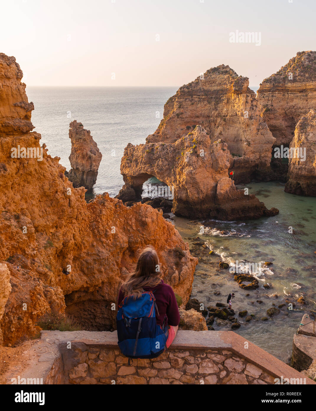 Weibliche Wanderer sitzt auf Mauer aus Stein und blickt über Felsen im Meer, Algarve, felsige Küste, Ponta da Piedade, Lagos, Portugal Stockfoto