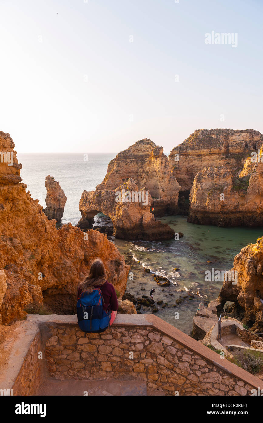 Weibliche Wanderer sitzt auf Mauer aus Stein und blickt über Felsen im Meer, Algarve, felsige Küste, Ponta da Piedade, Lagos, Portugal Stockfoto