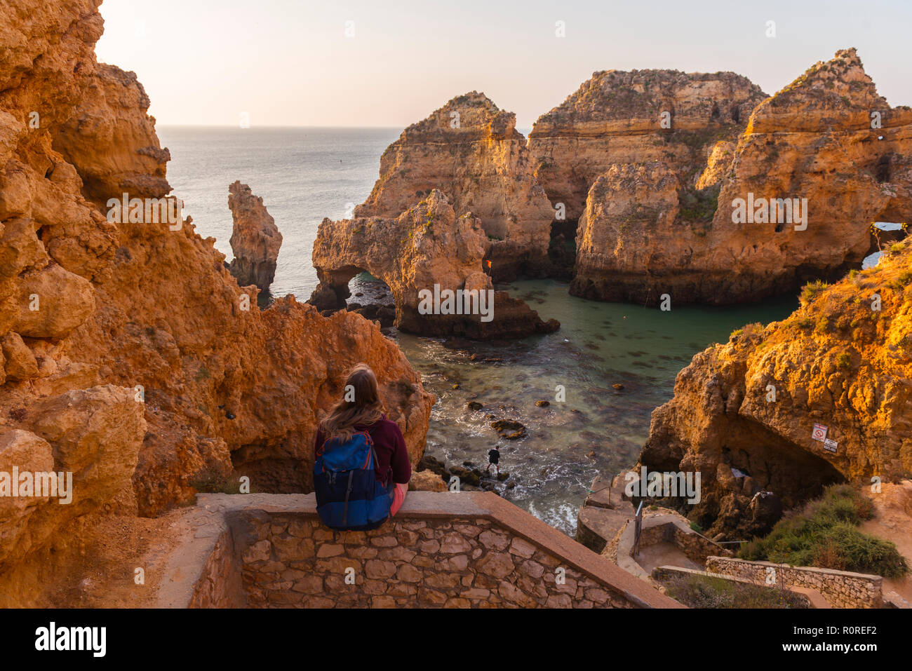 Weibliche Wanderer sitzt auf Mauer aus Stein und blickt über Felsen im Meer, Algarve, felsige Küste, Ponta da Piedade, Lagos, Portugal Stockfoto