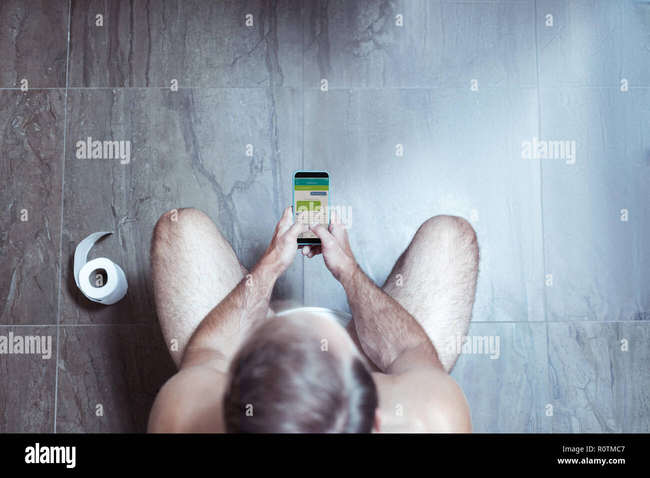 Blonde Mann mit seinem Smartphone während auf der Toilette sitzen Stockfoto