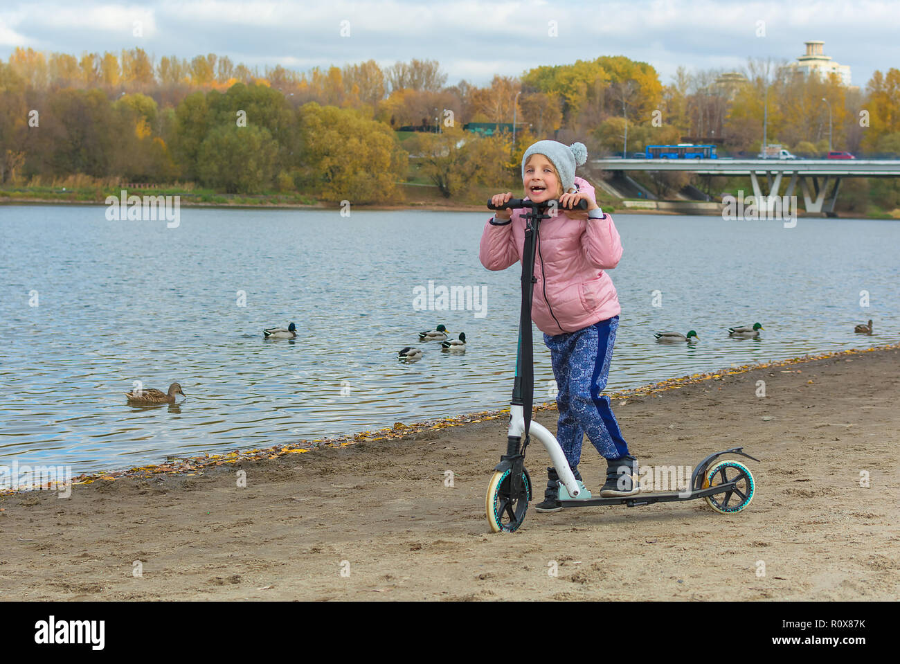 Herbst. Kleines Mädchen mit einem Scooter am Ufer des Flusses. Bewölkter Tag Stockfoto