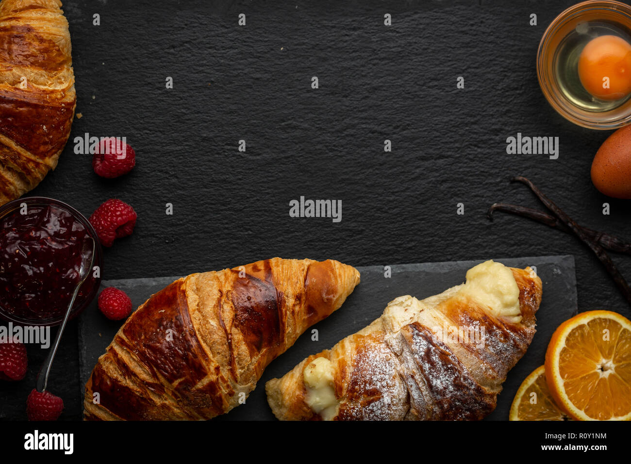 Croissants und Vanillepudding croissant umgeben mit Himbeeren, Himbeer Marmelade, Eier, Orangenscheiben und Vanilleschoten über Kopf geschossen Stockfoto