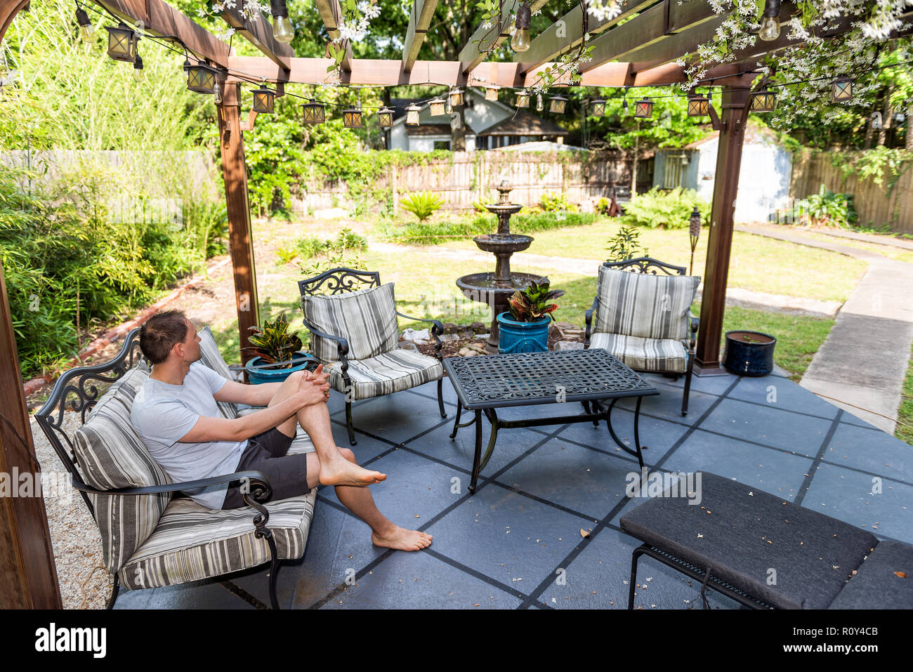 Junger Mann sitzen auf der Terrasse Lounge Chair in outdoor Frühling Blumen Garten im Hinterhof des Hauses zen mit Springbrunnen, Pergola Vordach Pavillon, Tisch, pla Stockfoto
