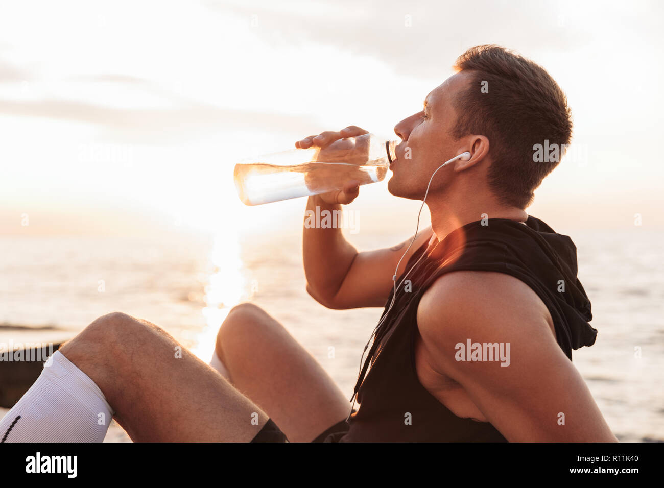 Bild von Stattlichen starken jungen Sportler im Freien am Strand Musik hören mit Kopfhörern Trinkwasser. Stockfoto