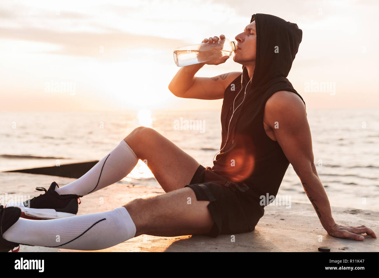 Bild von Stattlichen starken jungen Sportler im Freien am Strand Musik hören mit Kopfhörern Trinkwasser. Stockfoto
