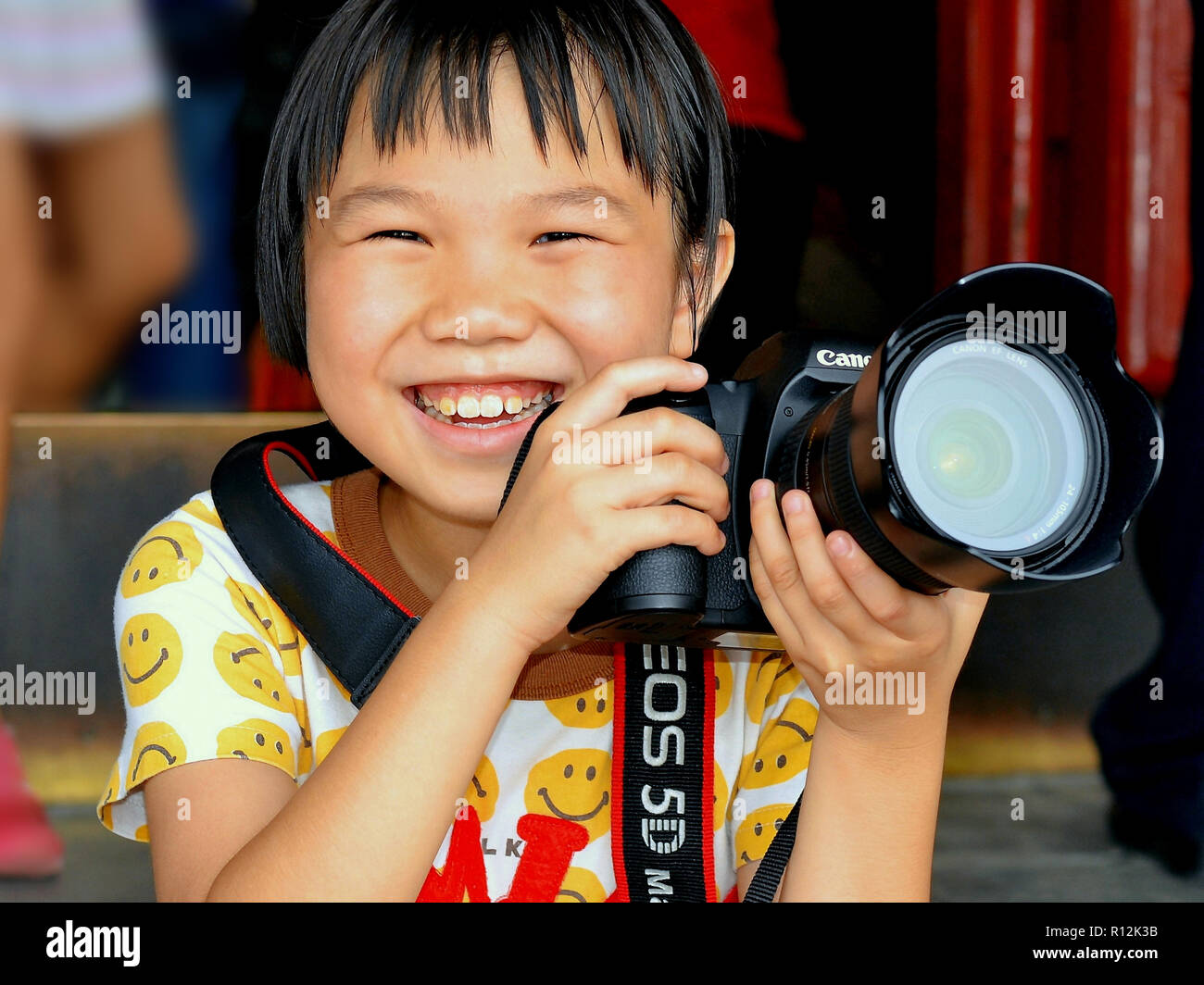 Kleine chinesische Mädchen mit einem Lächeln hält eine Canon EOS 5D mit einem Teleobjektiv in beiden Händen. Stockfoto