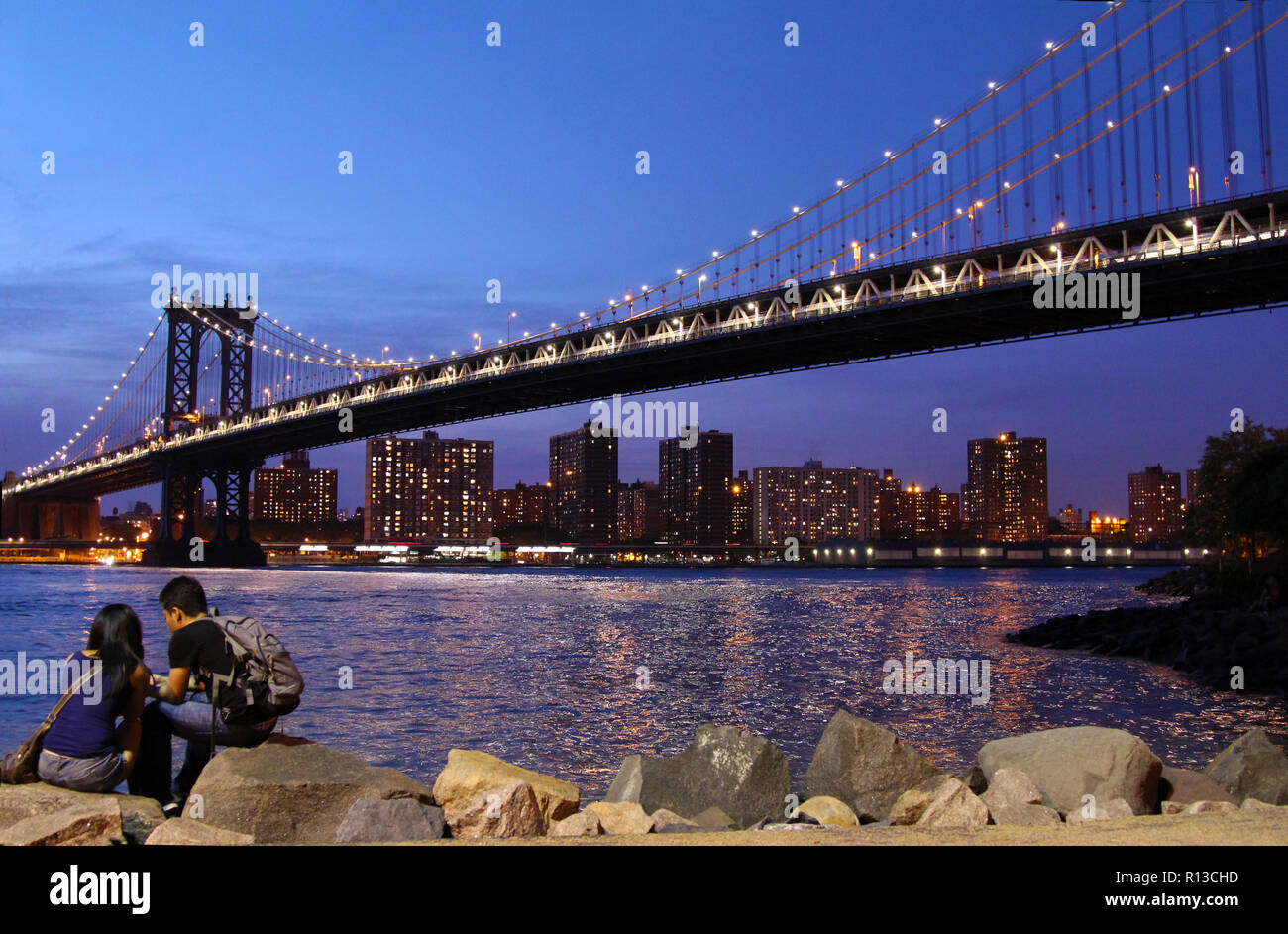 Jungen asiatischen Paar sitzt auf Felsen vor Manhattan Bridge ab, die im Rahmen der Manhattan Bridge Überführung (Dumbo), New York, NY gesehen Stockfoto