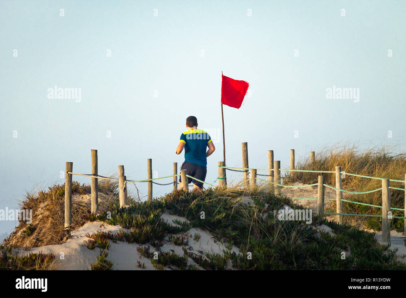 Junger Mann läuft auf der Promenade in der Nähe der Dünen. Red Flag. Blaues Hemd. Ansicht von hinten. Klaren Tag. Stockfoto