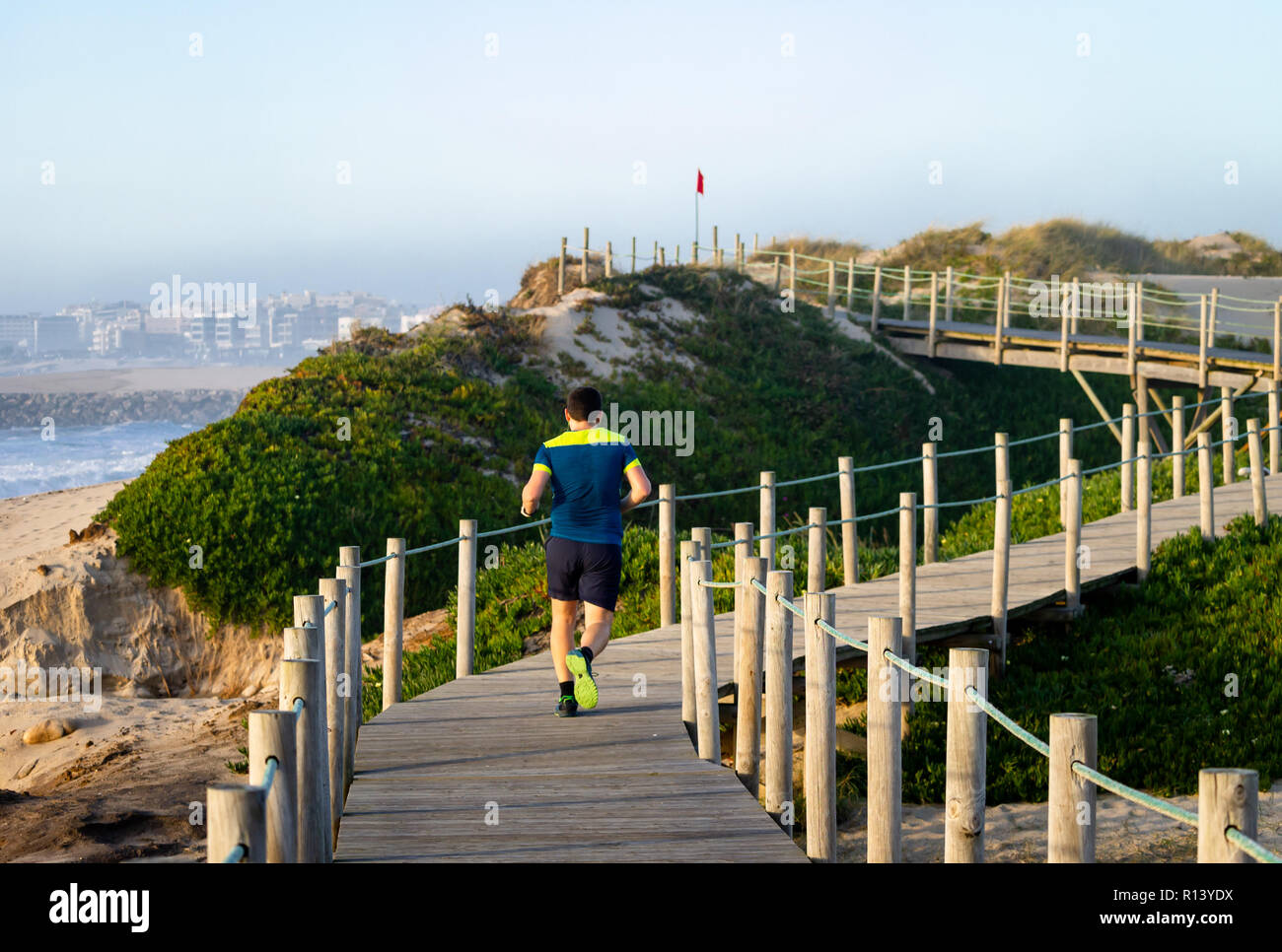 Junger Mann läuft auf der Promenade in der Nähe der Sand und das Meer in Richtung auf eine rote Fahne. Blaues Hemd. Ansicht von hinten. Klaren Tag. Stockfoto