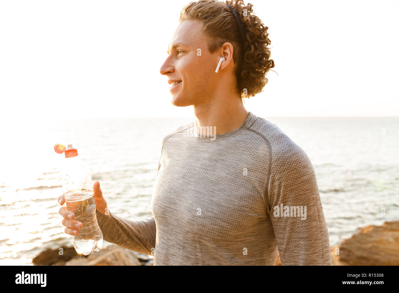 Foto von gutaussehenden jungen Sport Mann stand am Strand Trinkwasser Musik hören. Stockfoto