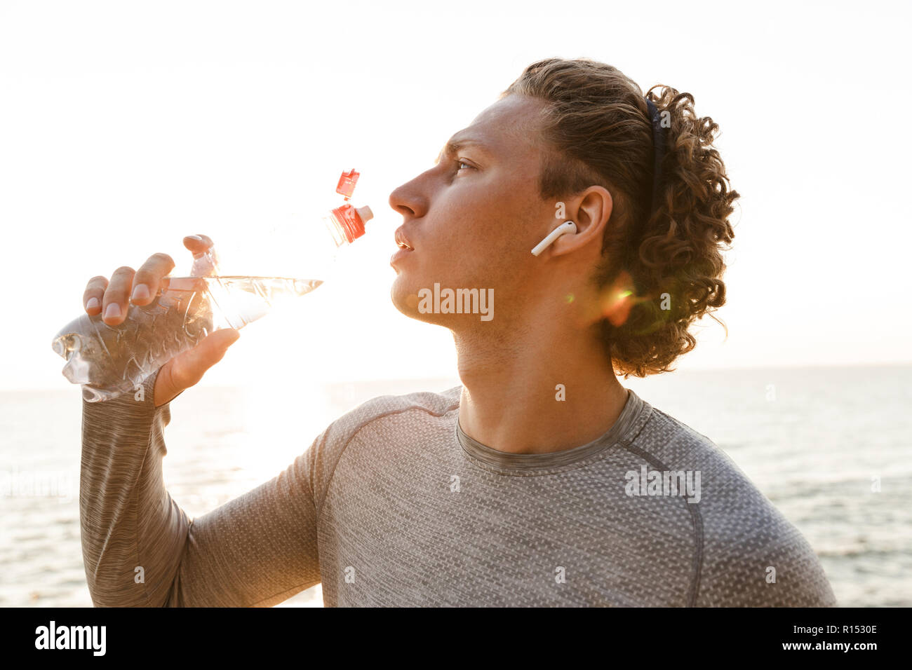 Foto von gutaussehenden jungen Sport Mann stand am Strand Trinkwasser Musik hören. Stockfoto