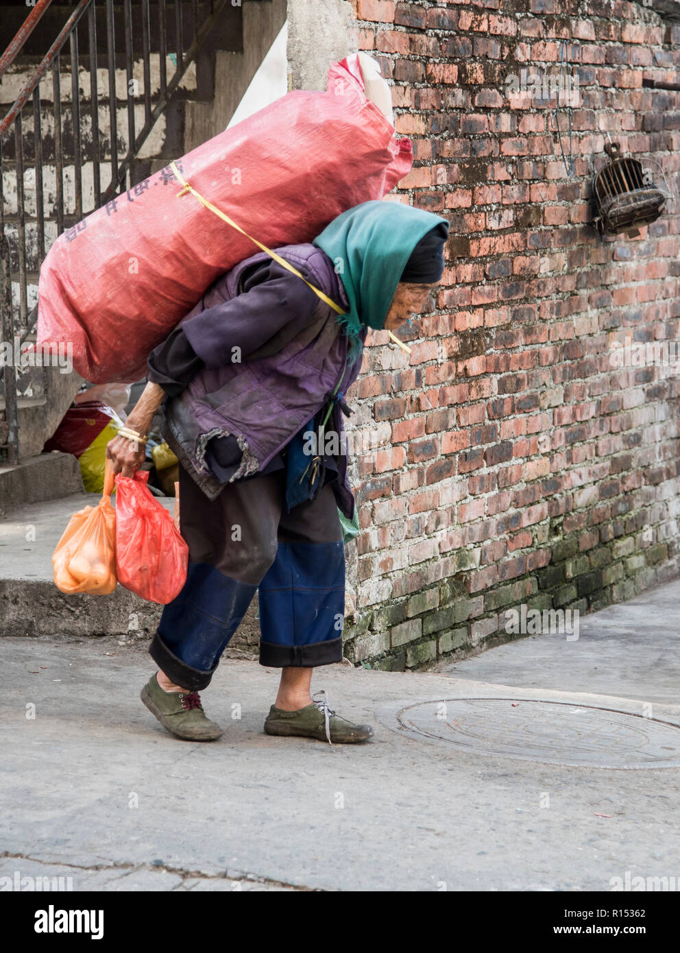 Eine alte chinesische Dame, die eine schwere Ladung trägt und es sehr anstrengend findet, zu Fuß zu gehen Stockfoto