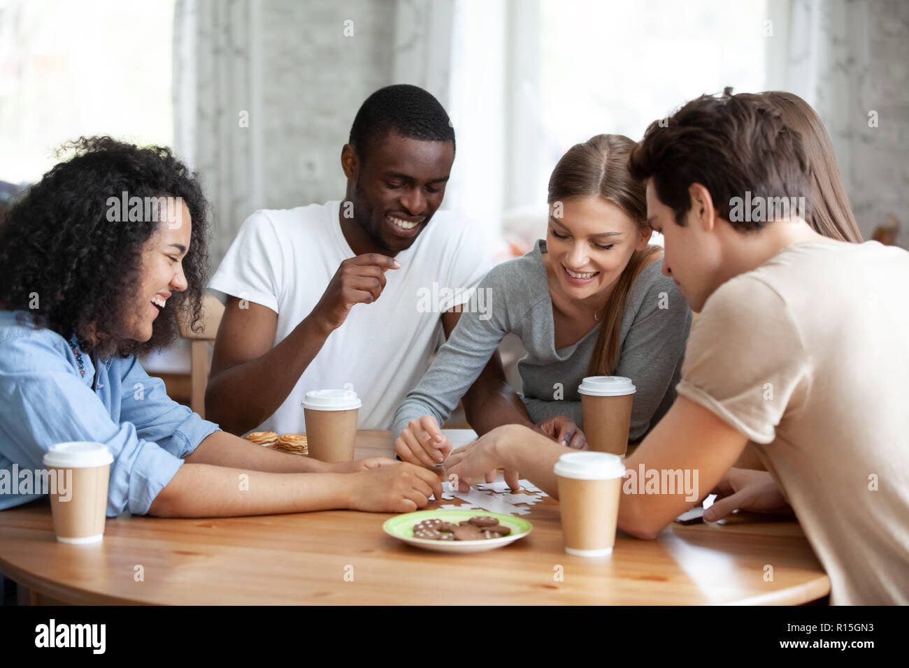 Diverse multirassischen Freunden fröhliche Mädchen und Jungs Kaffee trinken Spaß lachen Montage puzzle Puzzle zusammen am Tisch im Hause sitzen oder Stockfoto