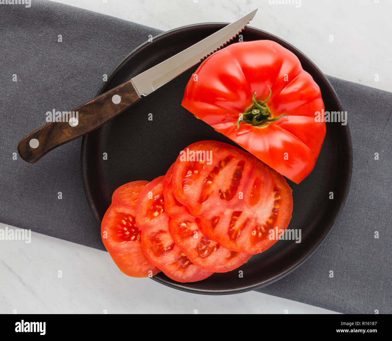 Große, saftige, rote Tomate teil geschnitten auf dunklen Teller mit Messer auf grauer Stoff, Stoff und weißem Marmor Hintergrund Stockfoto