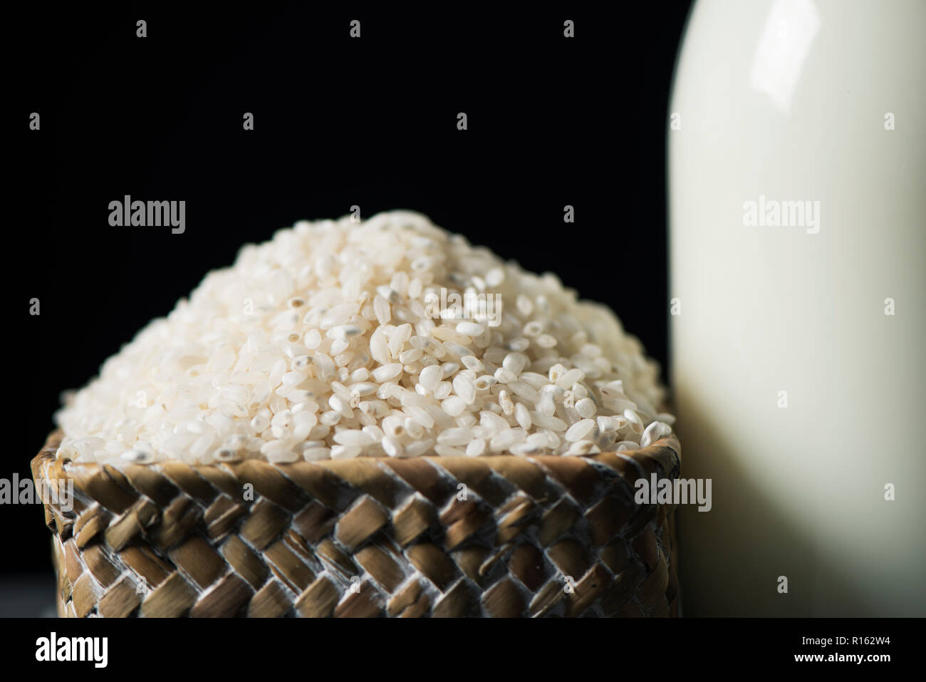 Nahaufnahme von einem Korb mit Reis und eine Glasflasche mit Reis, Milch, vor einem schwarzen Hintergrund Stockfoto