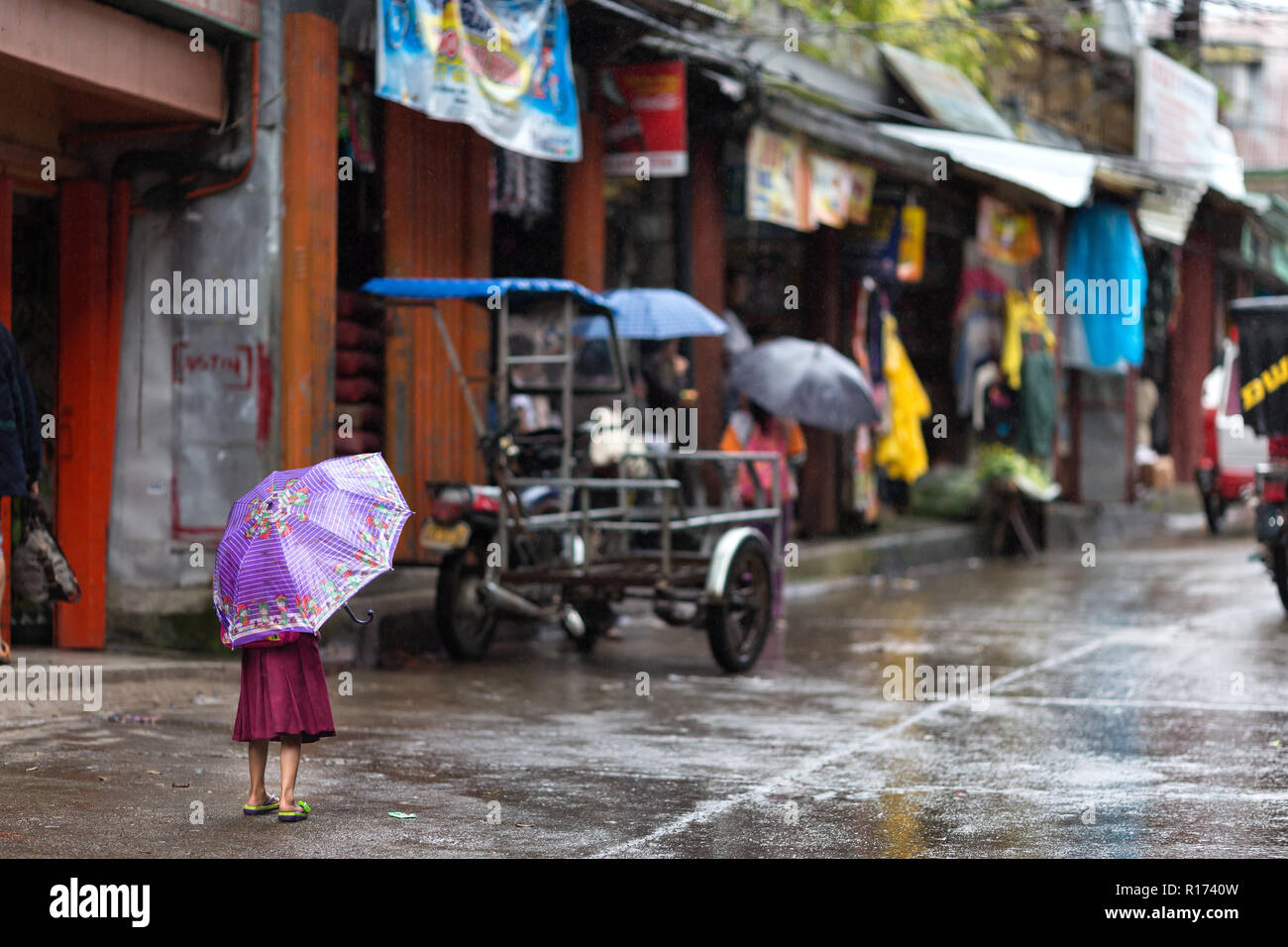 Süße kleine Mädchen Unterschlupf unter einem lila Schirm alleine stehen auf der Straße im Regen in Banuae Dorf, Philippinen gegenüber Menschen suchen Sie Stockfoto