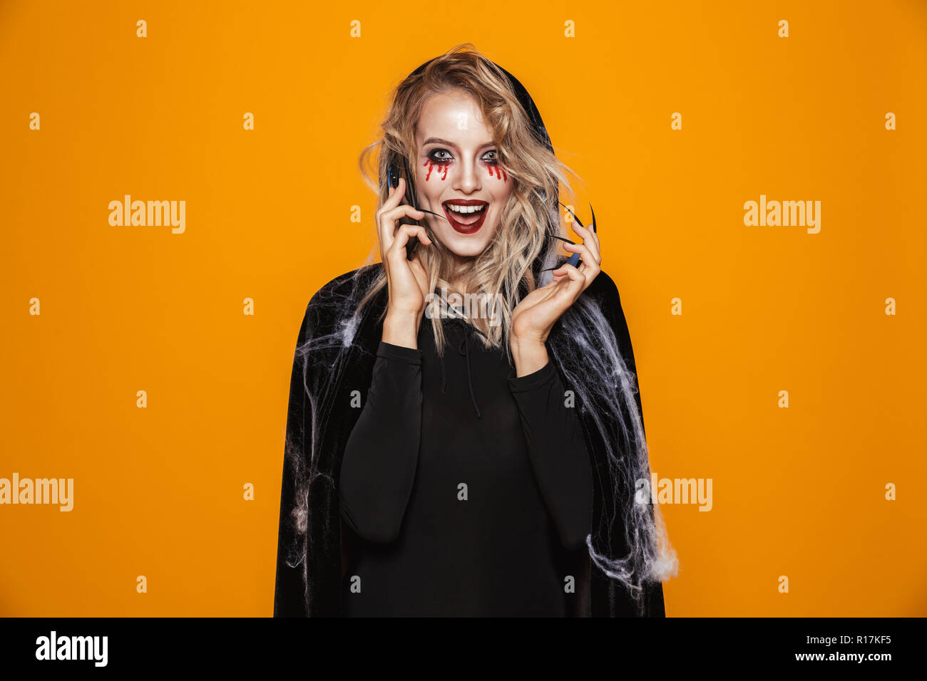Lächelnde blonde Frau Hexe mit Make-up sprechen von Smartphone und suchen Kamera über orange isoliert Stockfoto