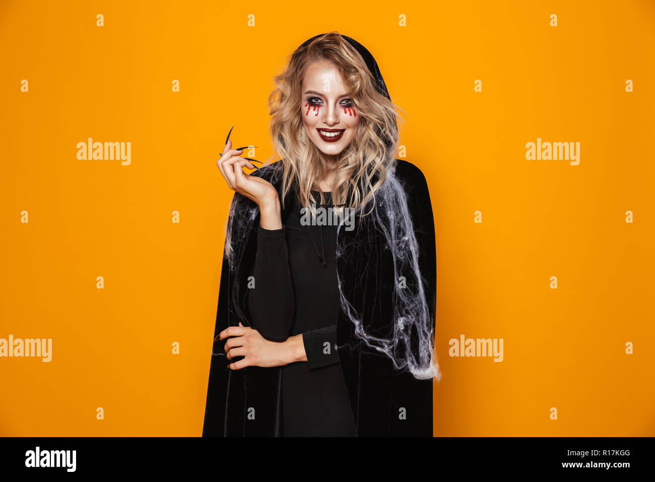 Lächelnde blonde furchtsame Frau Hexe in schwarzen Mantel suchen Kamera über orange isoliert Stockfoto