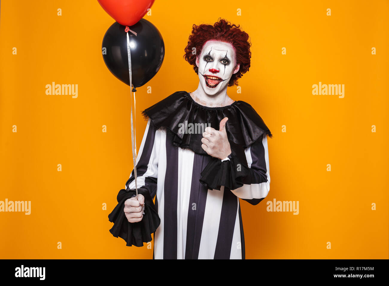 Zornigen Mann in Scary clown Halloween Kostüm holding Ballons über Gelb Hintergrund isoliert gekleidet, mit Daumen hoch Stockfoto