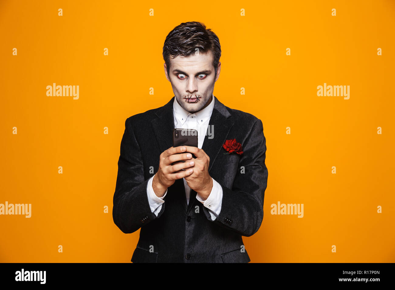 Schockiert zombie Mann Bräutigam wih Halloween Make-up mit Smartphone über orange isoliert Stockfoto