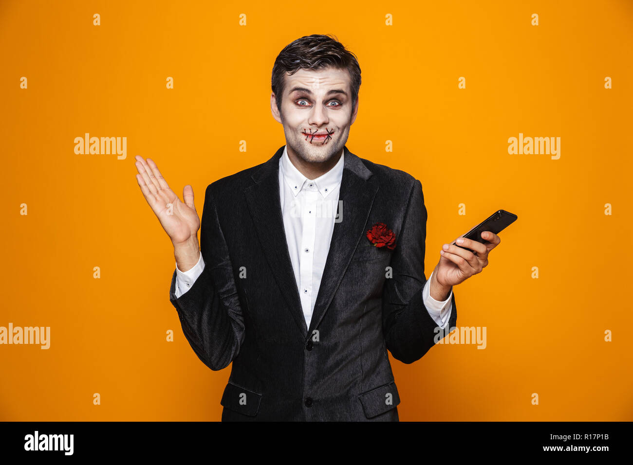 Schöne fröhliche Mann Zombie mit Halloween Make-up Holding Smartphone und zur Kamera über orange isoliert lächelnd Stockfoto