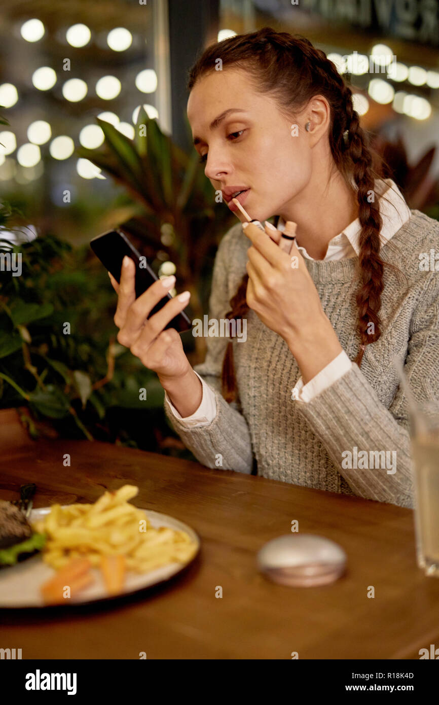 Ziemlich Casual Mädchen Anwendung Lip Gloss nach dem Essen während der Bildschirm des Smartphones im Cafe suchen Stockfoto