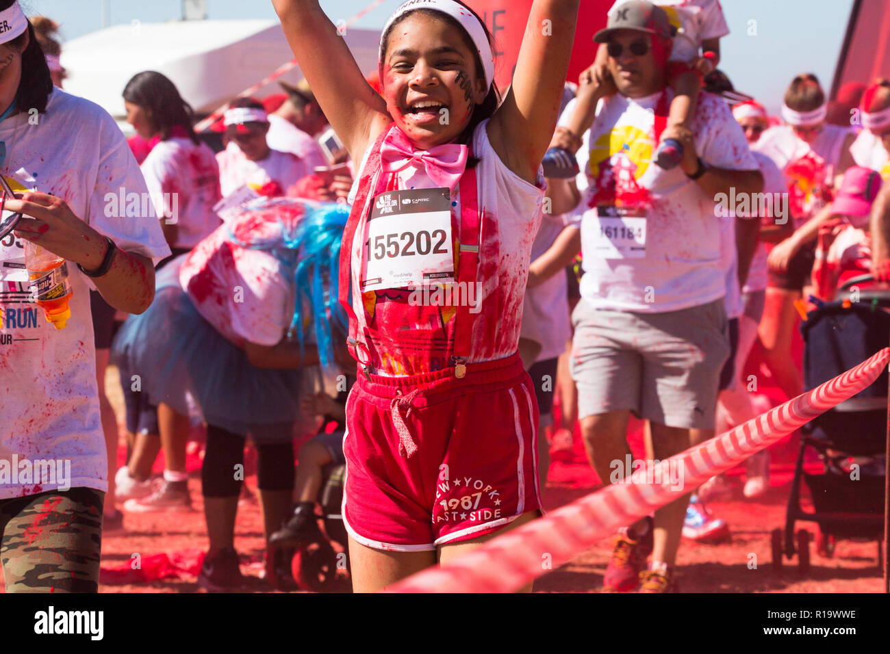Kleines Mädchen von gemischten Rennen oder farbige Ethnie ca. 8-10 Jahre alt springen auf und ab, die Arme in der Luft in roter Farbe pulverbeschichtet, während ein Sommer Colour run Veranstaltung angehoben Stockfoto
