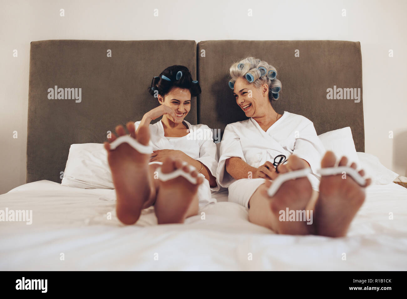Glückliche Mutter und Tochter sitzen am Bett mit Curling Rollen fixiert auf das Haar. Zwei Frauen sitzen auf dem Bett entspannen beim Pflegen Haar und Füße. Stockfoto